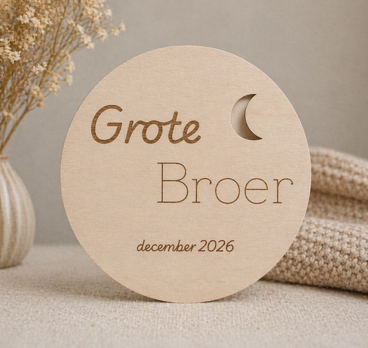 Ronde houten schijf met “Grote Broer”, maantje en datum, op een warme neutrale achtergrond.
