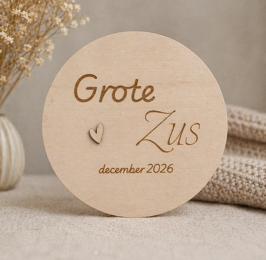 Ronde houten schijf met “Grote Zus”, hartje en datum, op een warme neutrale achtergrond.